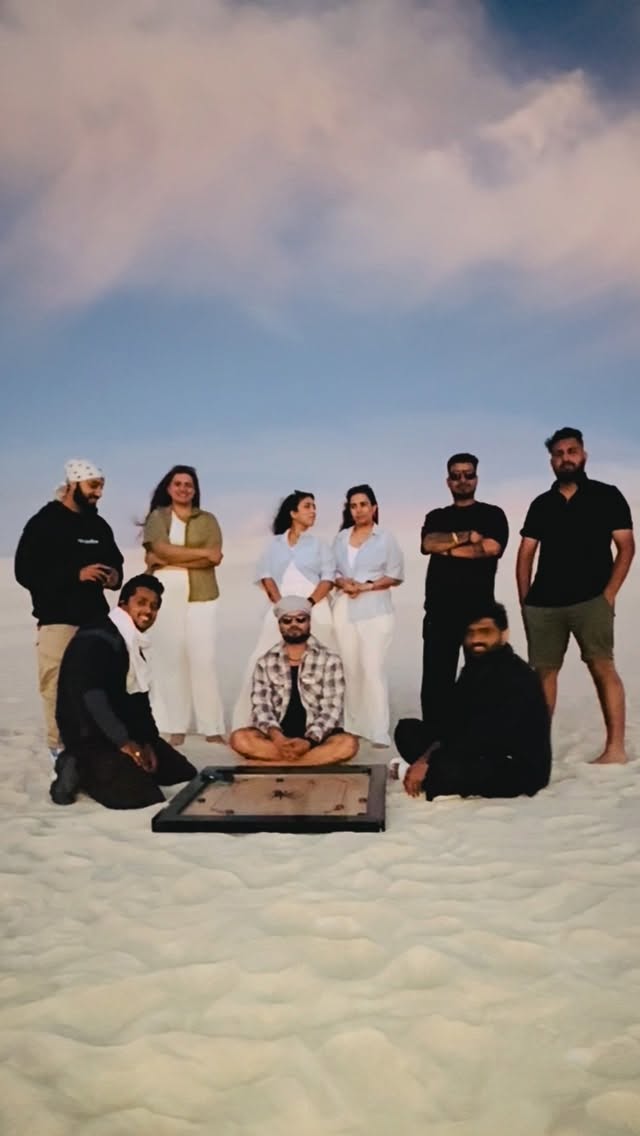 A group standing and sitting around a wooden carrom board on pale sand under a cloudy dusk sky
