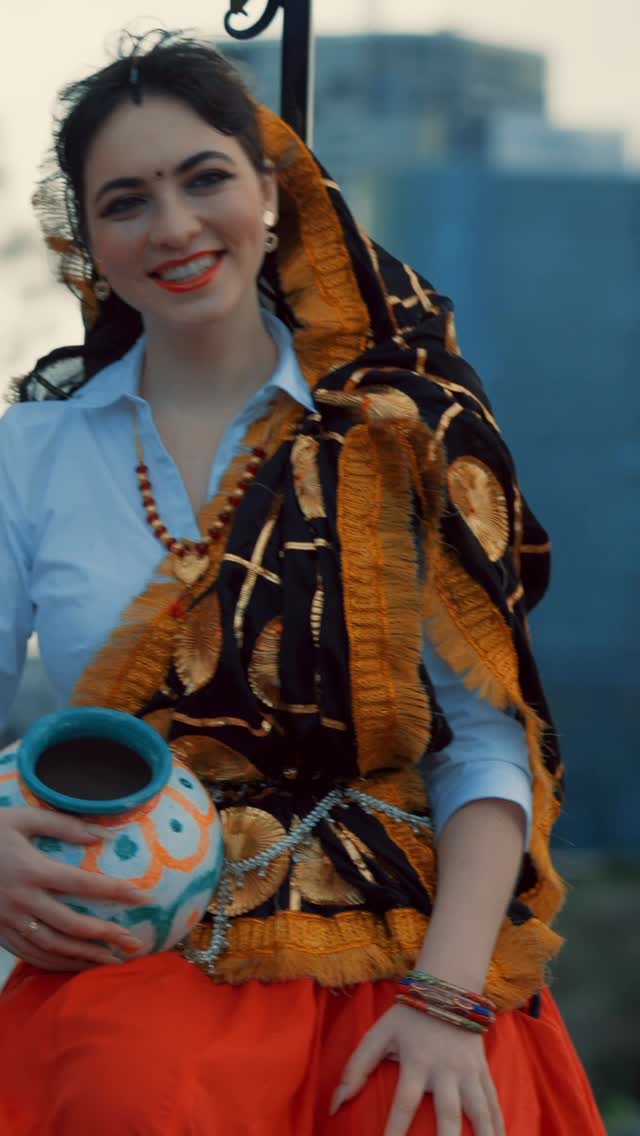 A smiling woman in a black and gold embroidered shawl and orange skirt holding a painted clay pot