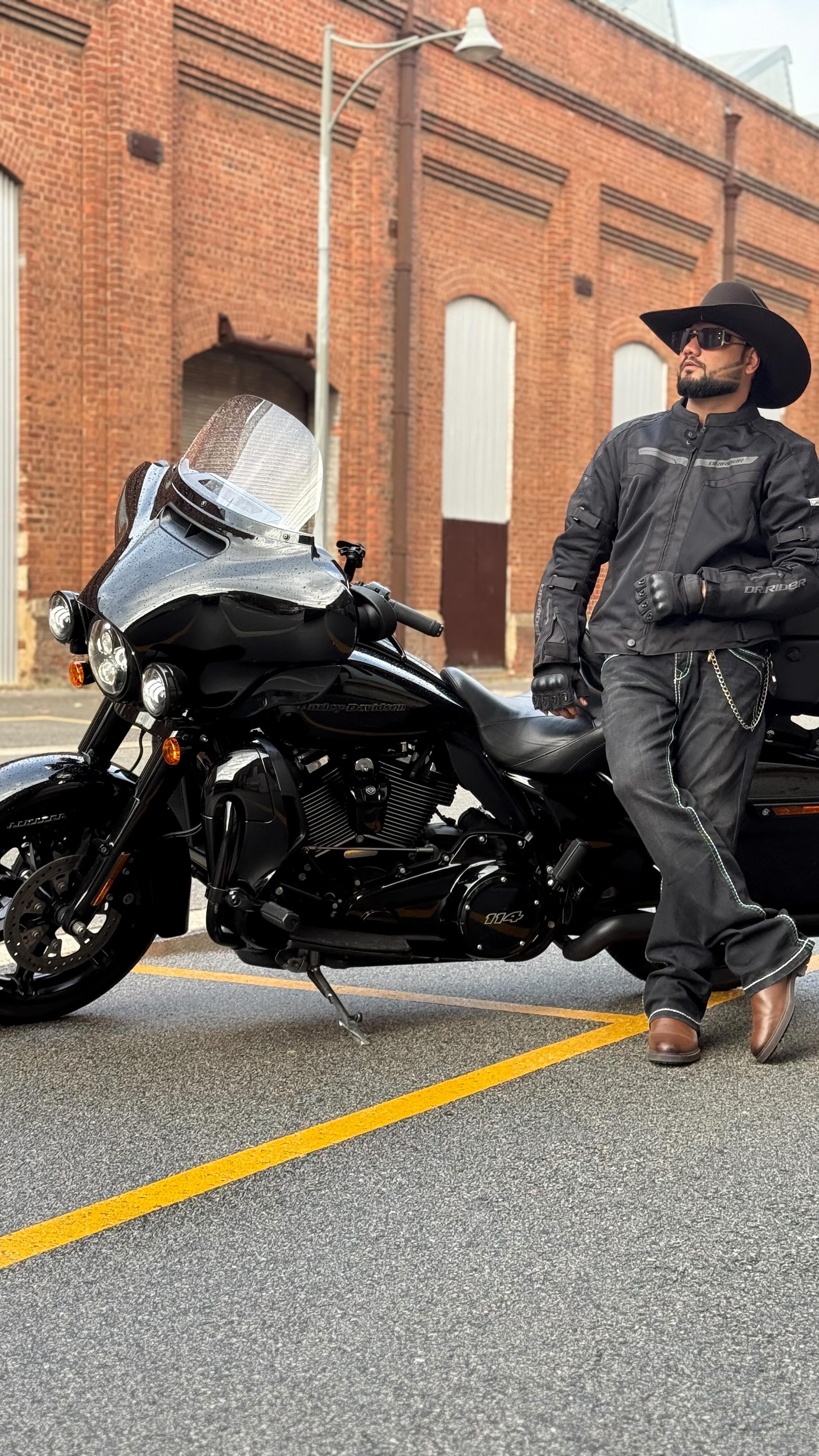 A man in a cowboy hat and riding jacket leaning on a black motorcycle in front of a red brick building